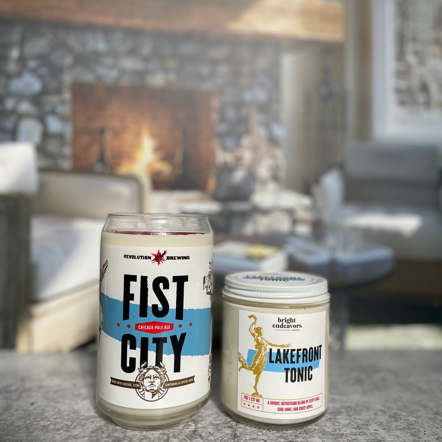 Two candles with blue accent labels on a table with a fireplace and couch in the background.